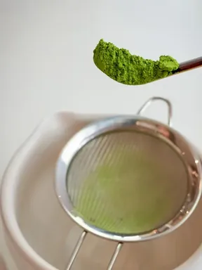A spoonful of matcha held over a fine sieve and a white bowl